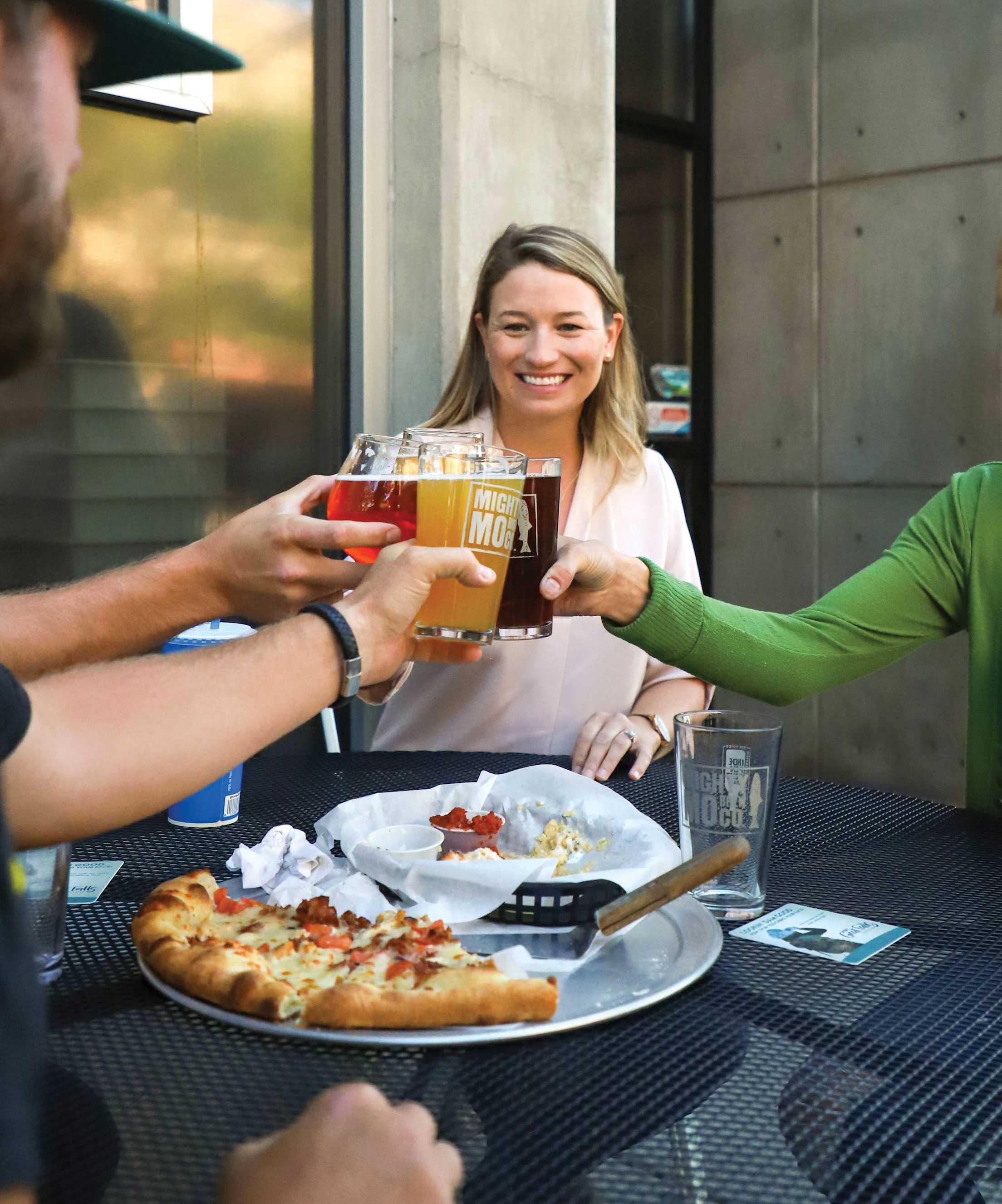 three people cheers with pint glasses of beer over a pizza