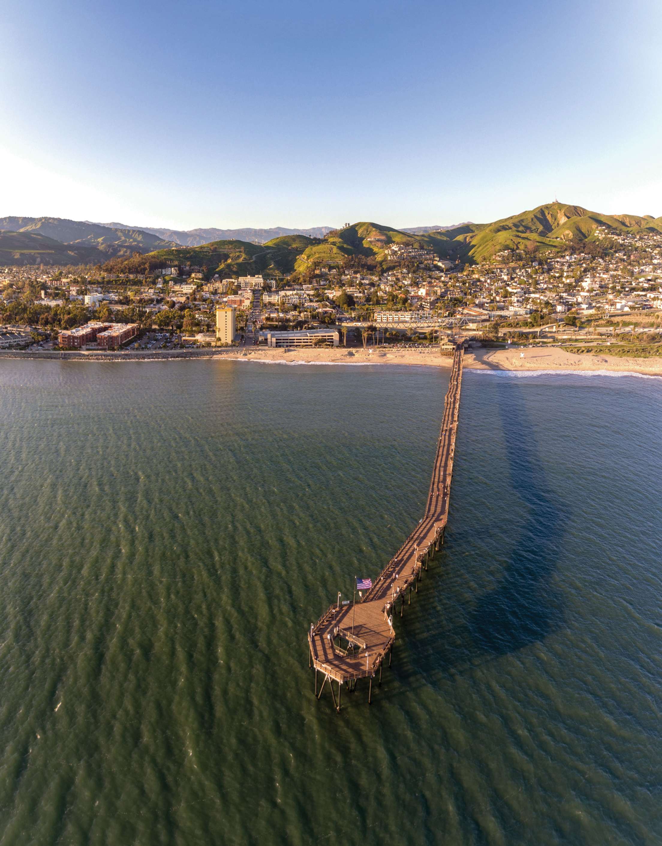 a long pier stretches out over the water in front of a beach and green hills