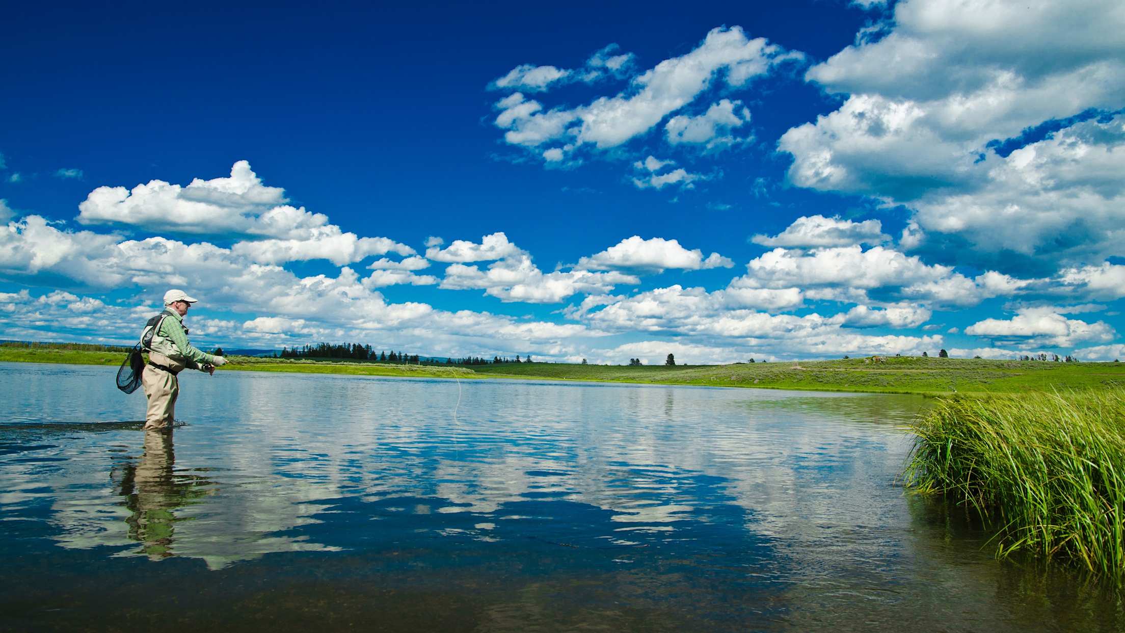 an older white man fly fishes in a blue lake with a bright blue sky