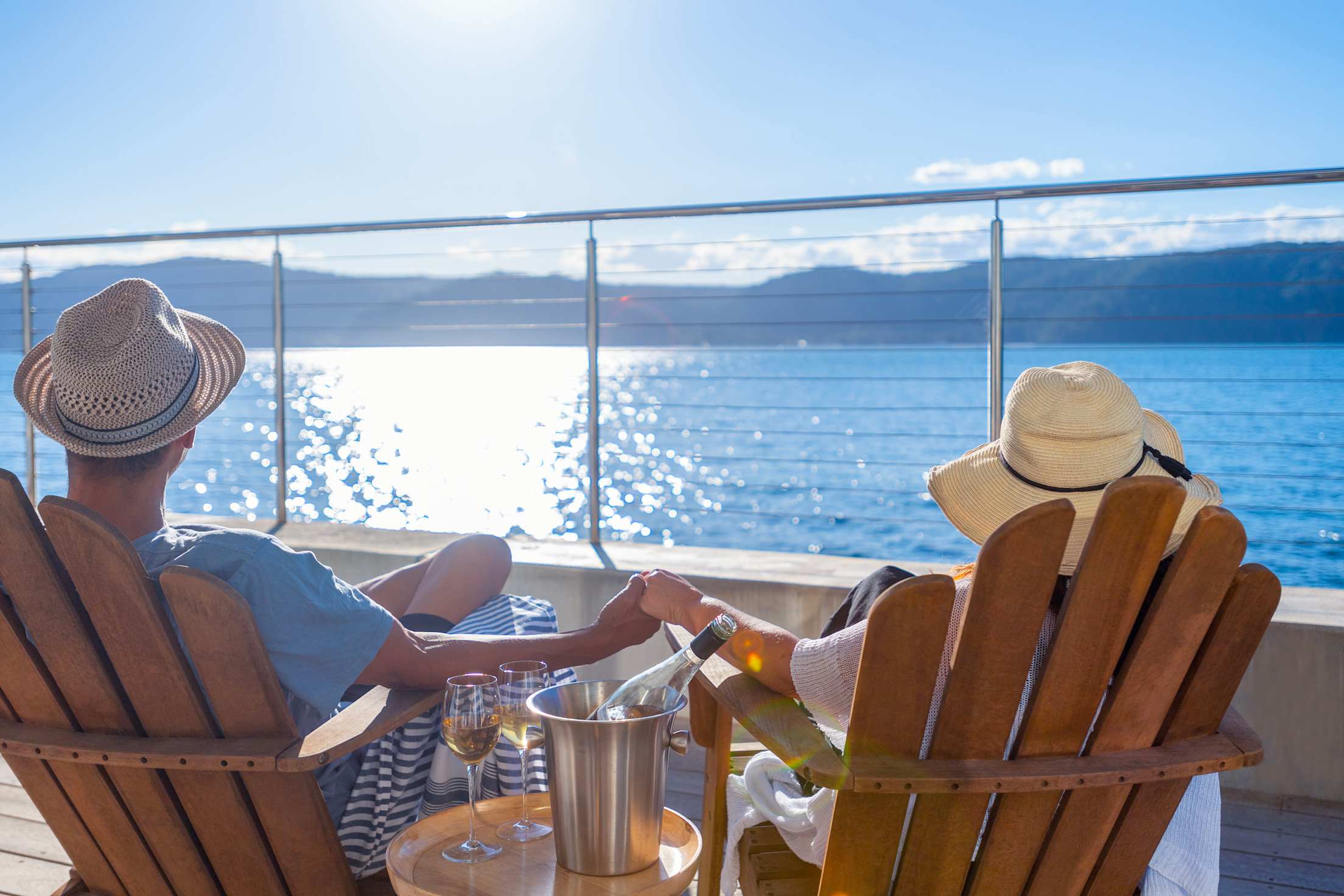 a couple seen from the back sits on a cruise ship deck in lounge chairs with glasses of wine