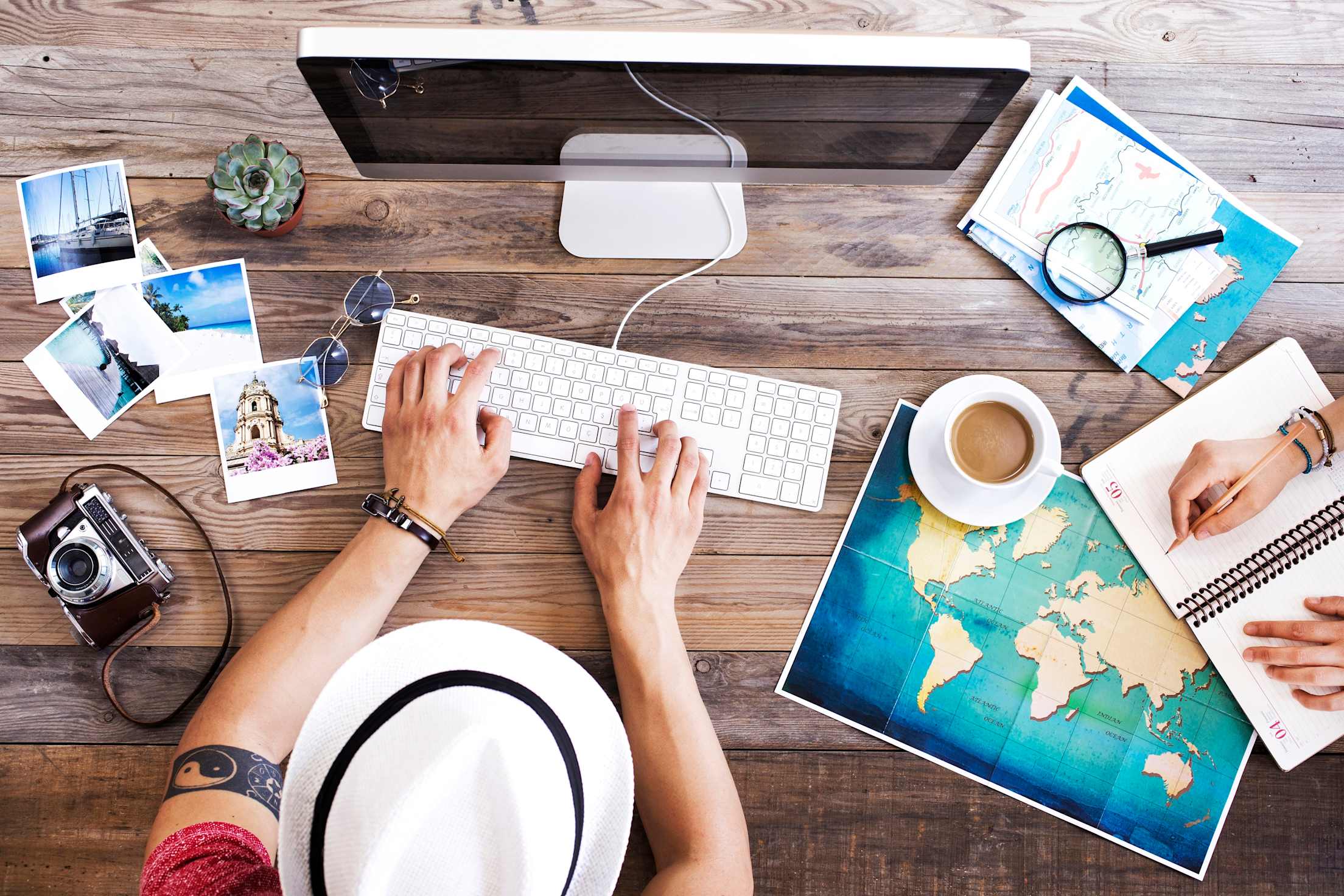 an overhead shot of a person with a hat and maps spread out on a table sitting at a computer planning a trip