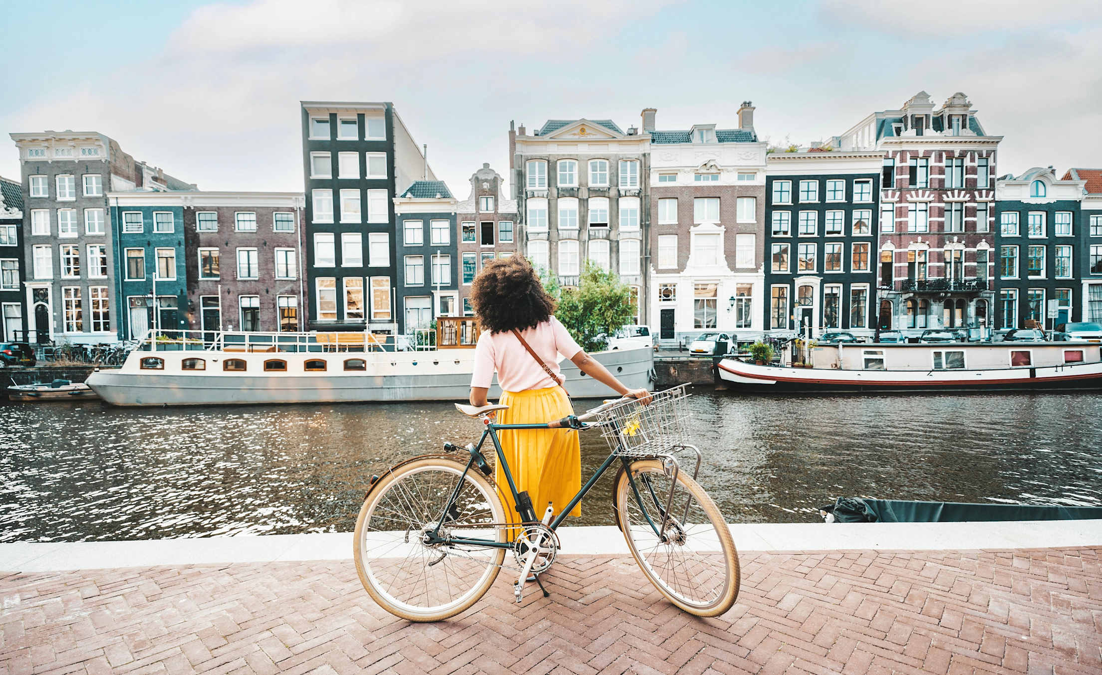 a woman in a yellow skirt faces away from the camera and toward a canal in Amsterdam, leaning on a bicycle
