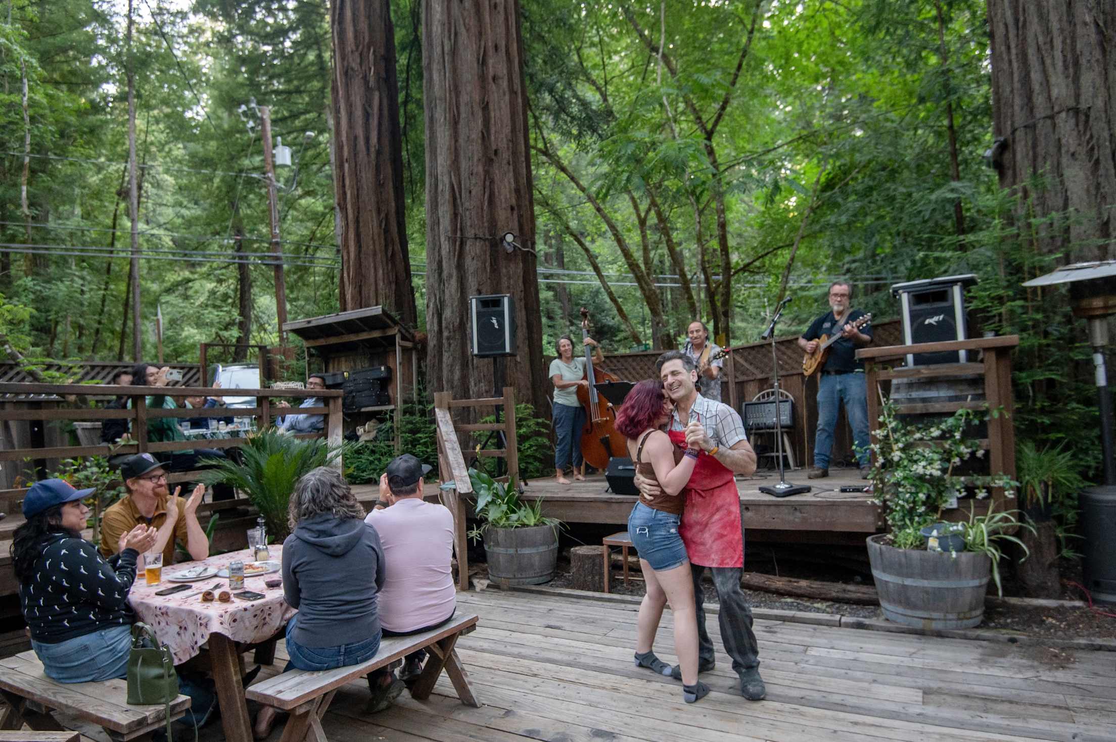two people dance on a wooden patio to live music surrounded by tall redwood trees