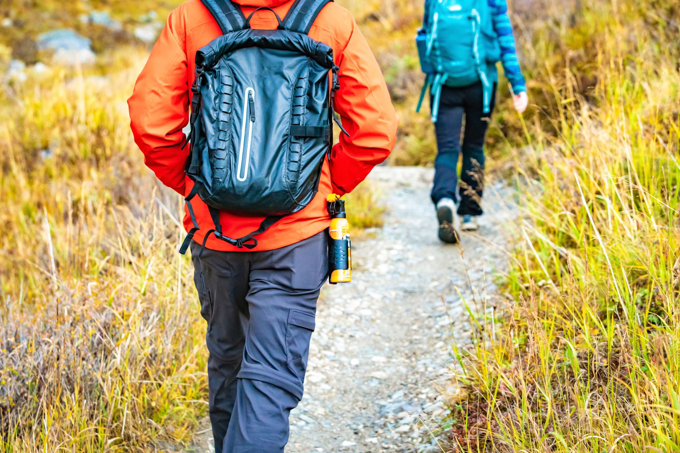 two people hike on a trail, one carrying a yellow canister of bear spray