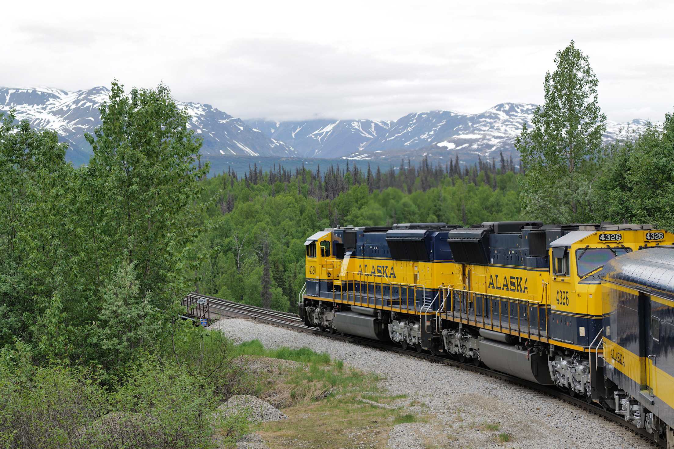 a yellow train that reads 'alaska' on the side runs by snow-capped mountains
