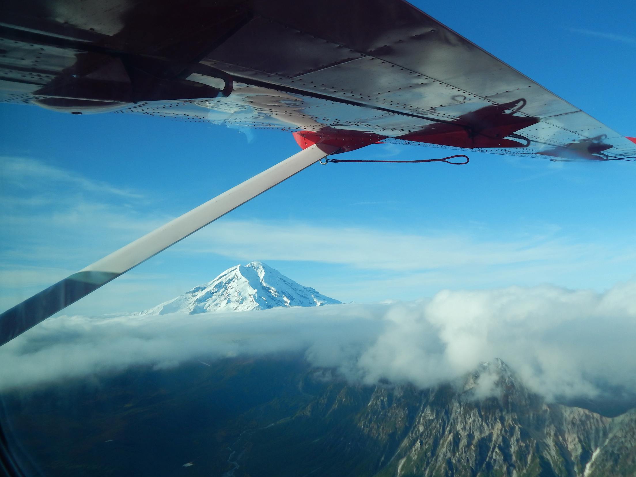 a snow-capped mountain as seen from a small plane, with its wing in the foreground