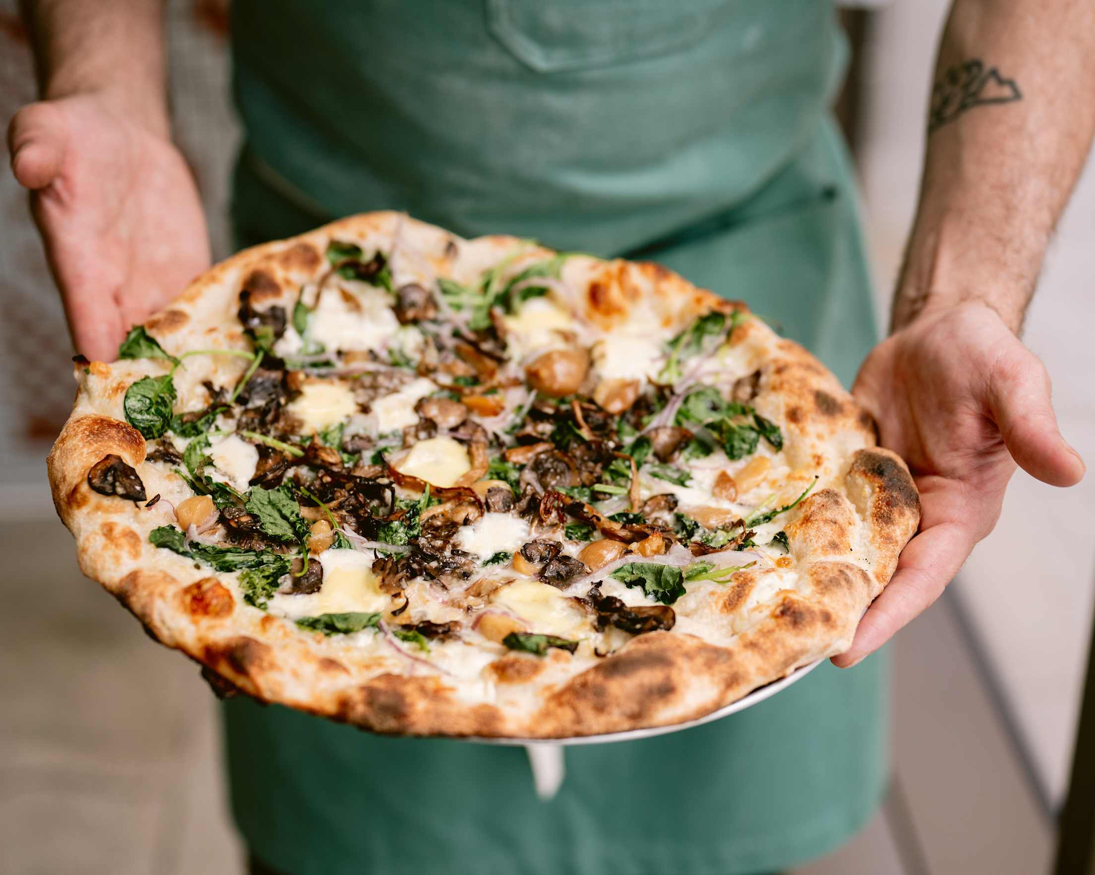 a pizza with mushrooms on it is held a by a person in a green apron