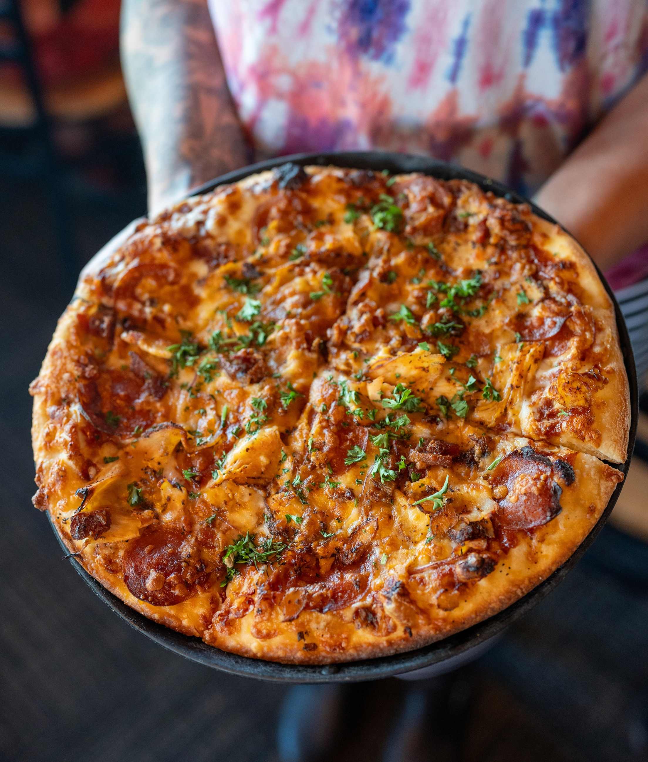 a person in a tie-dye shirt holds up a pizza
