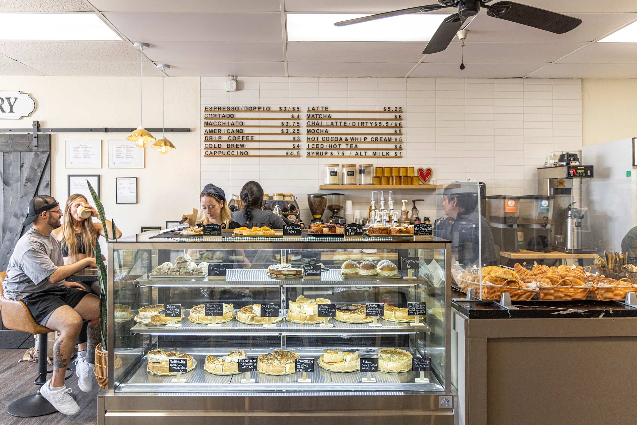 two people sit at a pie counter drinking coffee and smiling while two people are seen working behind the counter