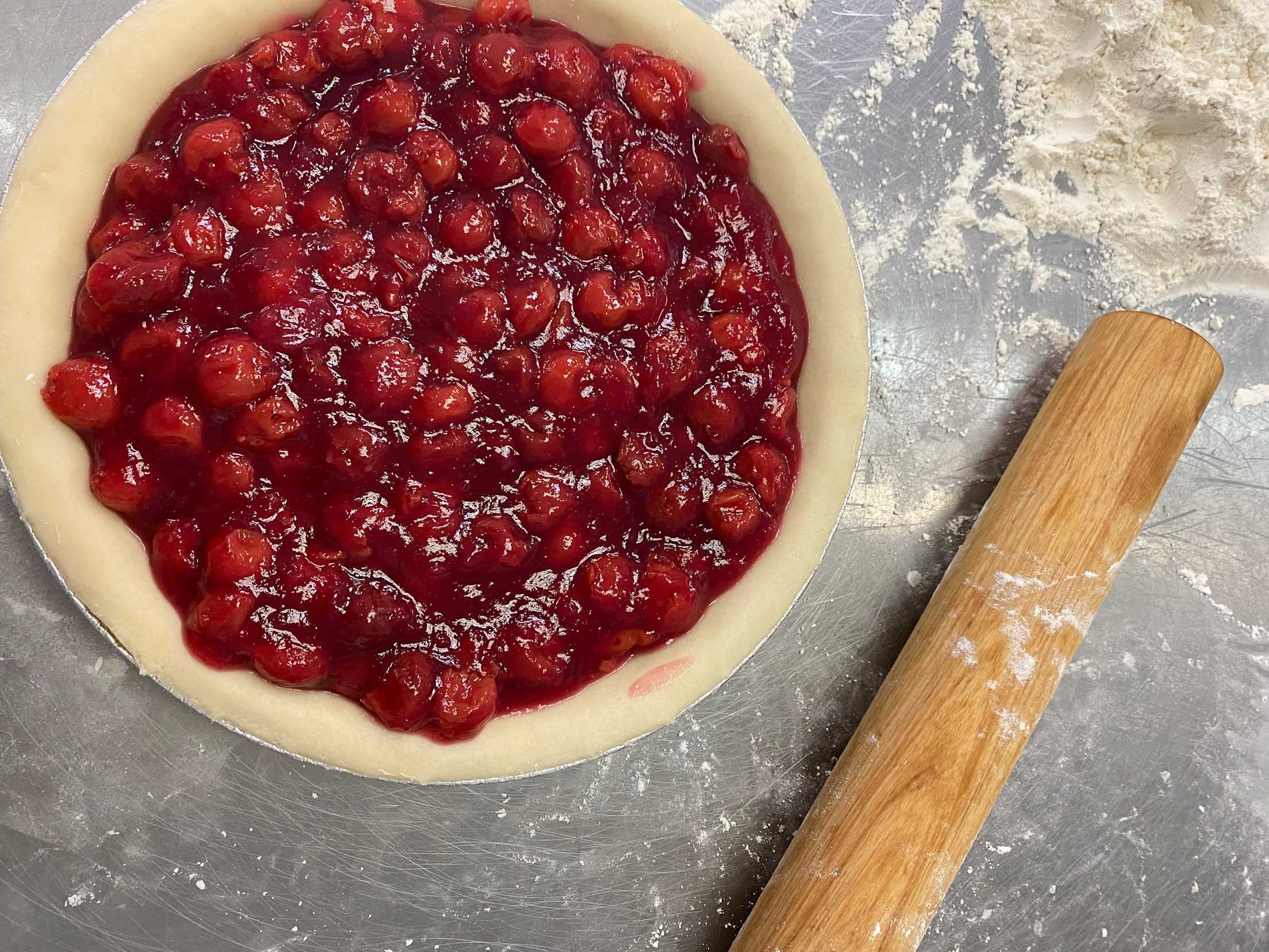 a cherry pie without a top crust next to a rolling pin and some flour