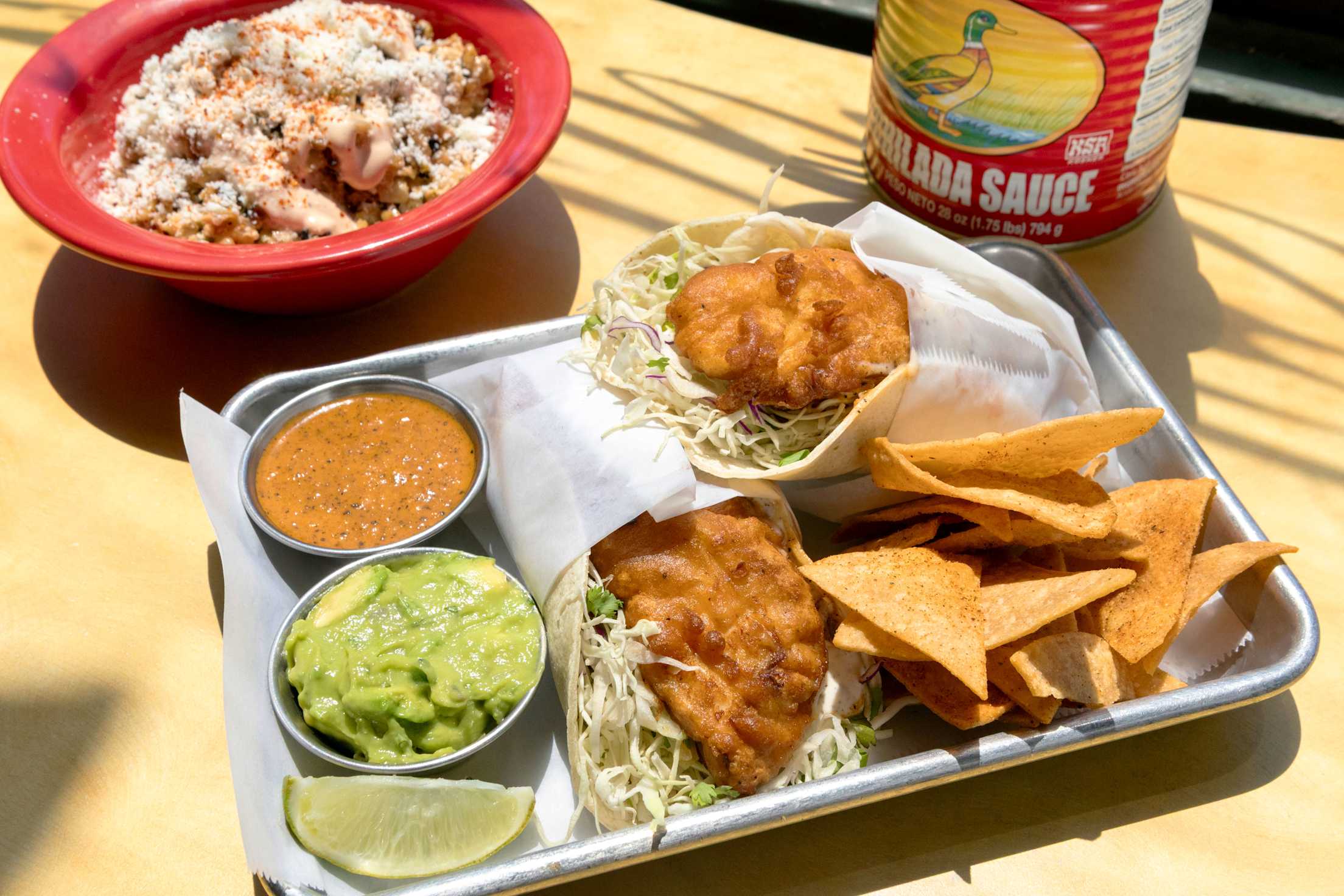 A tray of fish tacos with guacamole, salsa, and tortilla chips, plus a bowl of elote on the side.