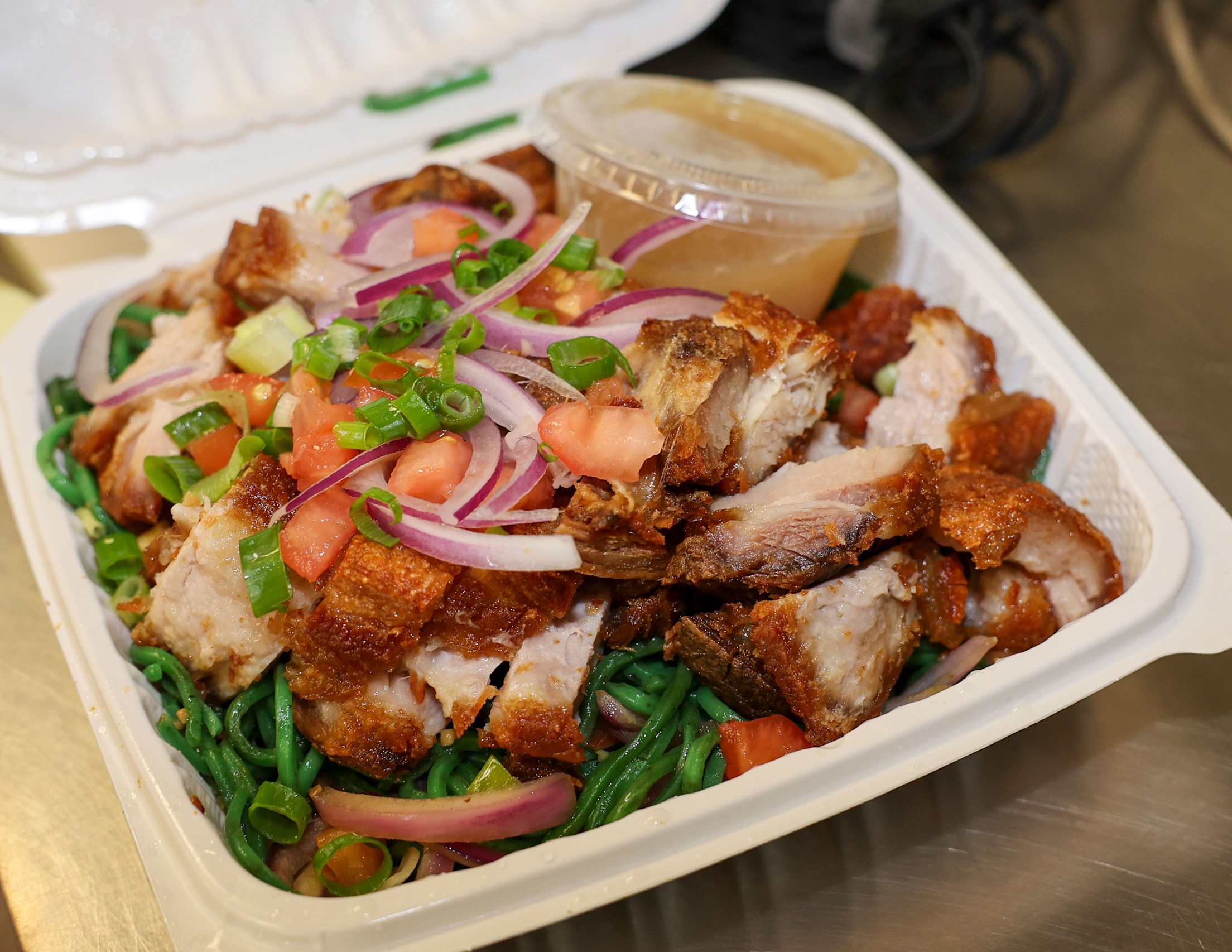 A takeout box of roasted pork belly over moringa noodles, topped with sliced vegetables.