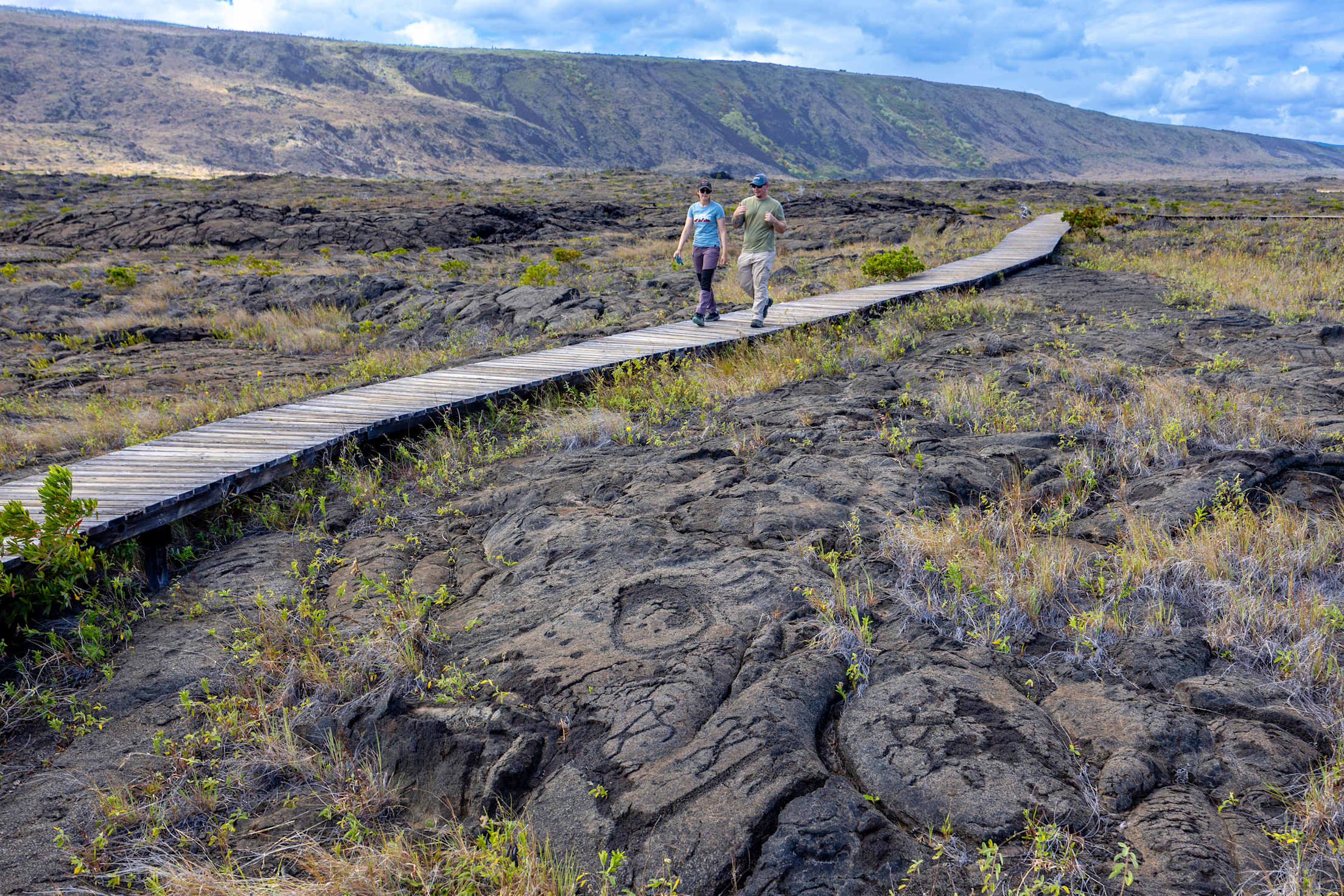Two people walk along a wooden boardwalk with petroglyphs visible in hardened lava flow