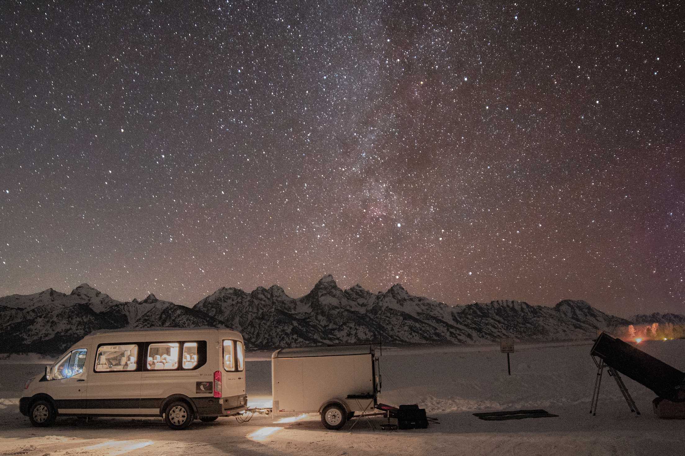 thousands of stars are seen above mountains with a camper van and telescope lit up in the foreground