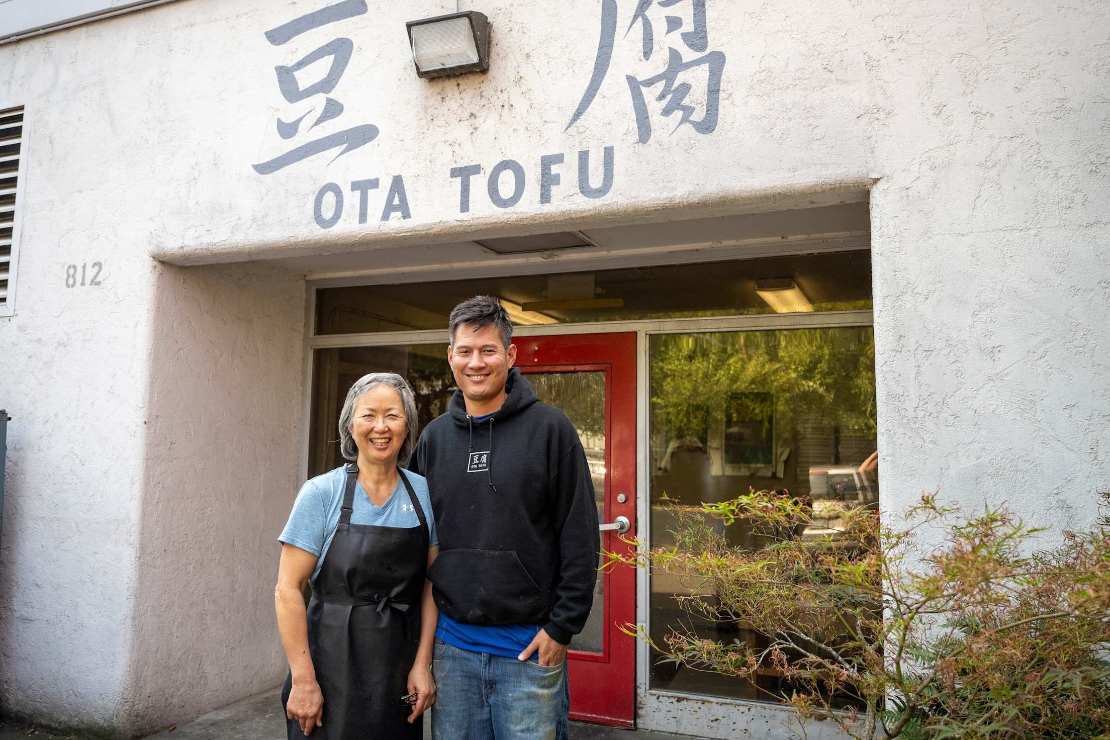 an older Japanese American woman and her adult son stand in front of a storefront that reads 'Ota Tofu' in English and Japanese