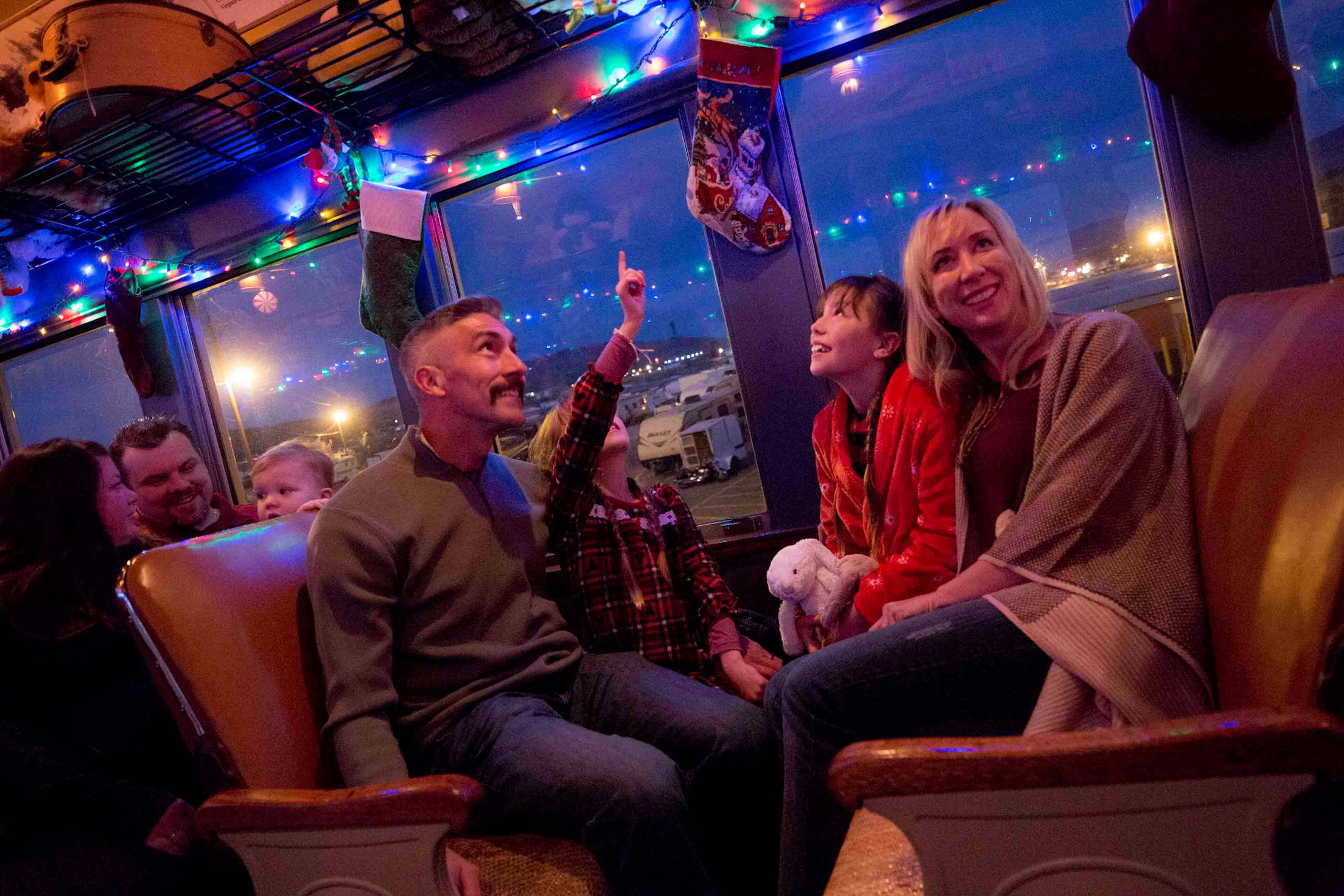 a family of four sits on a festively lit train, with a kid pointing at the Christmas lights on the ceiling