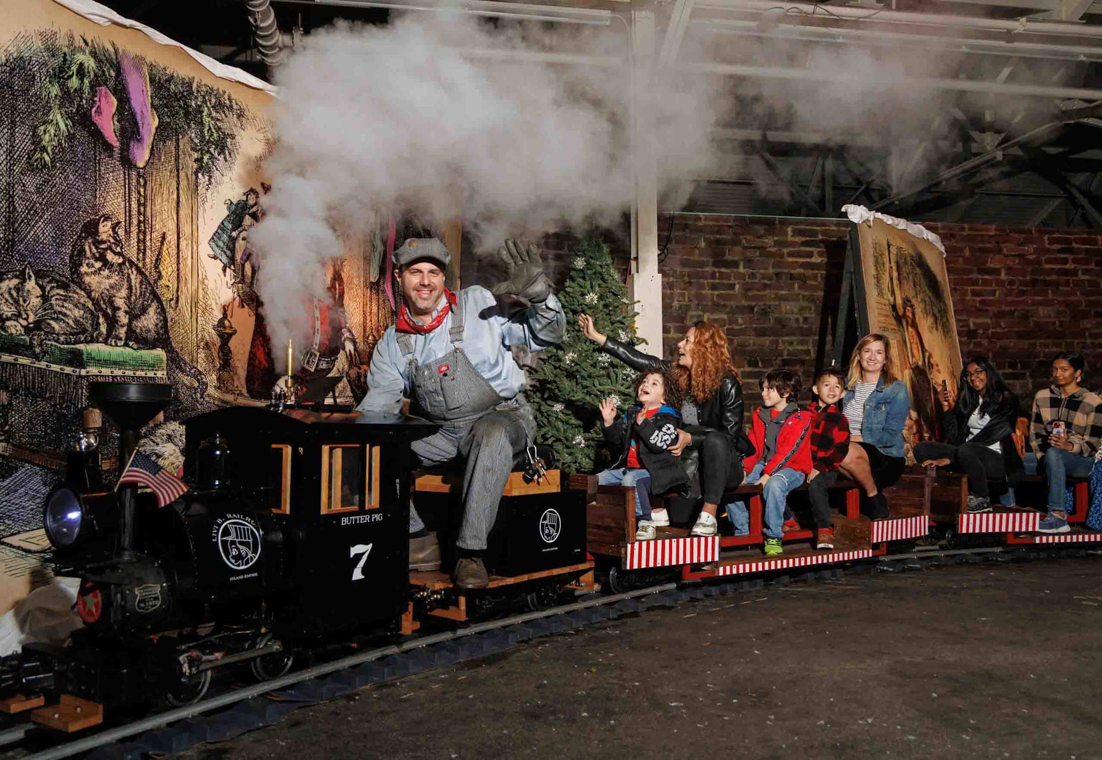 a driver smiles while driving a miniature steam train full of smiling kids through a Christmas decoration-filled room