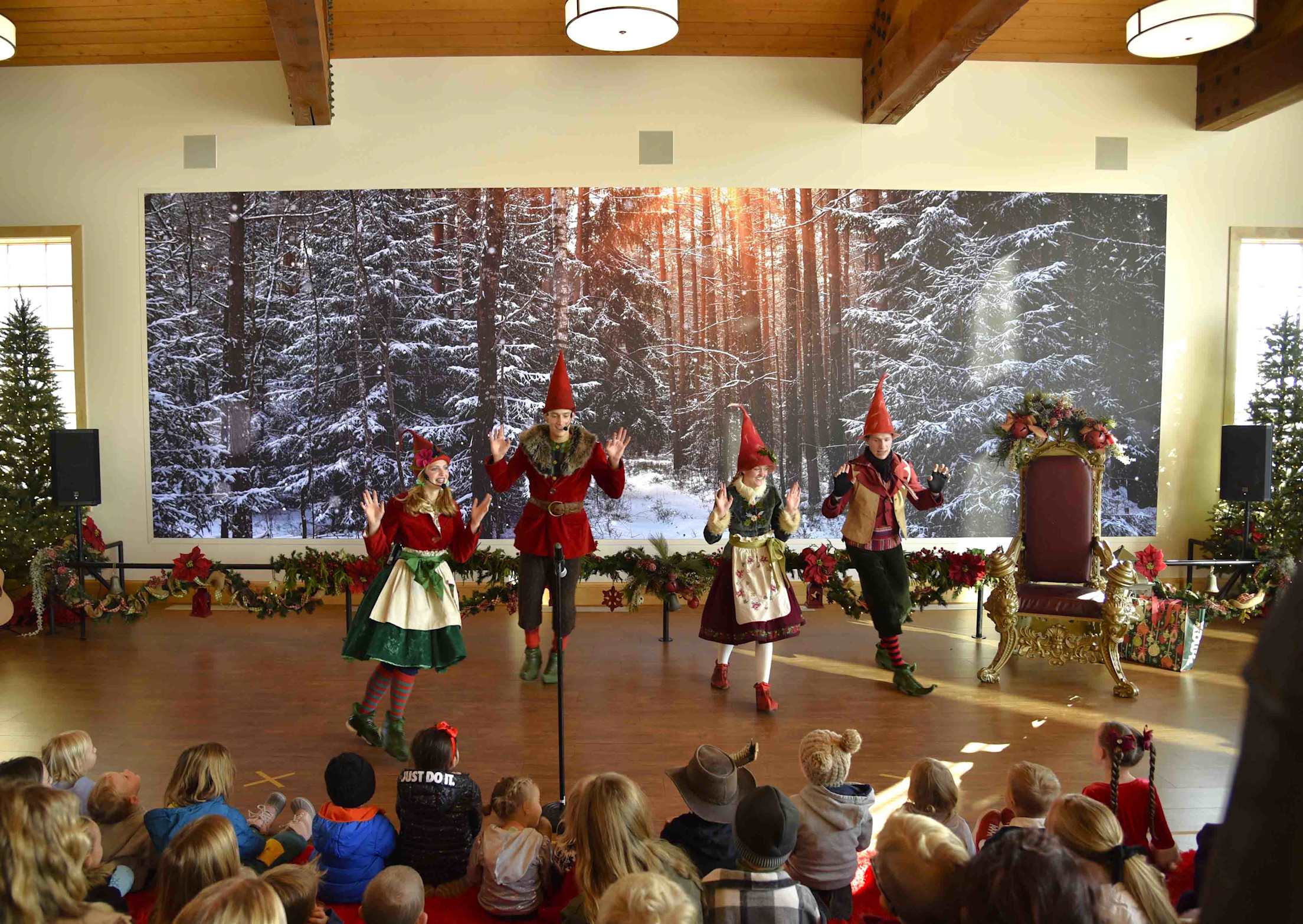 children seated in a room watch a live performance with four people dressed as elves