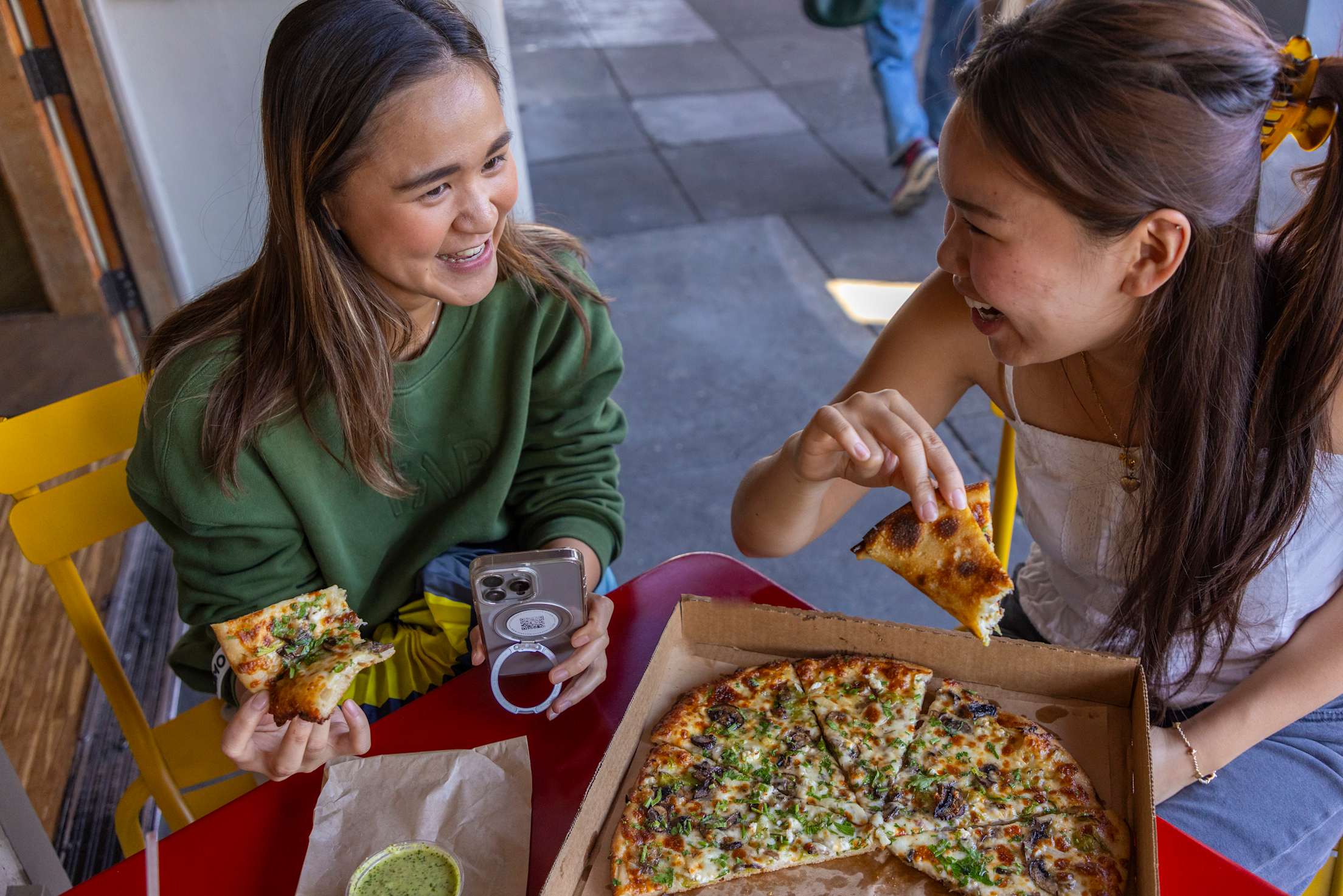 two young women sit eating pizza outside
