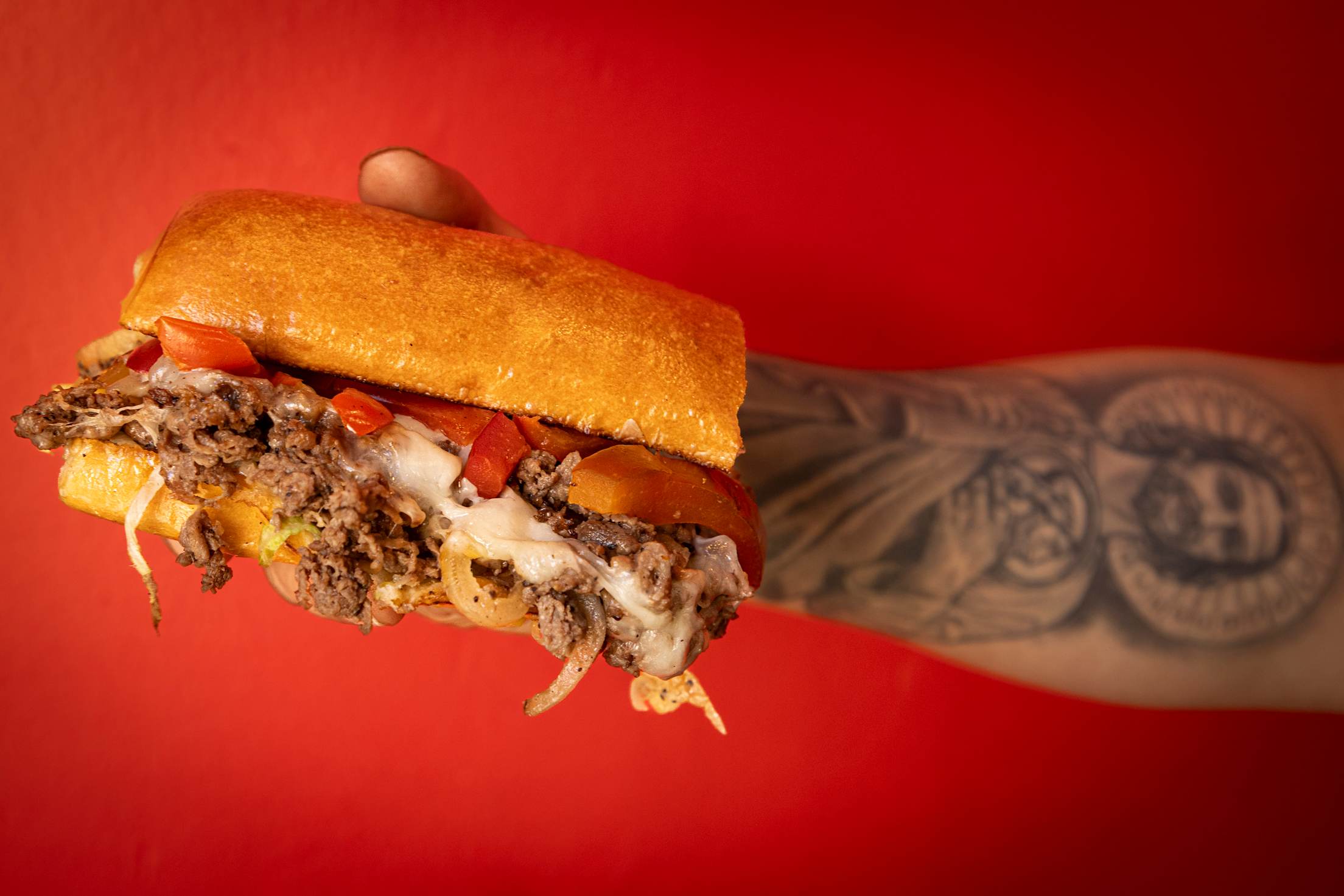 a tattooed arm holding a loaded philly cheesesteak sandwich