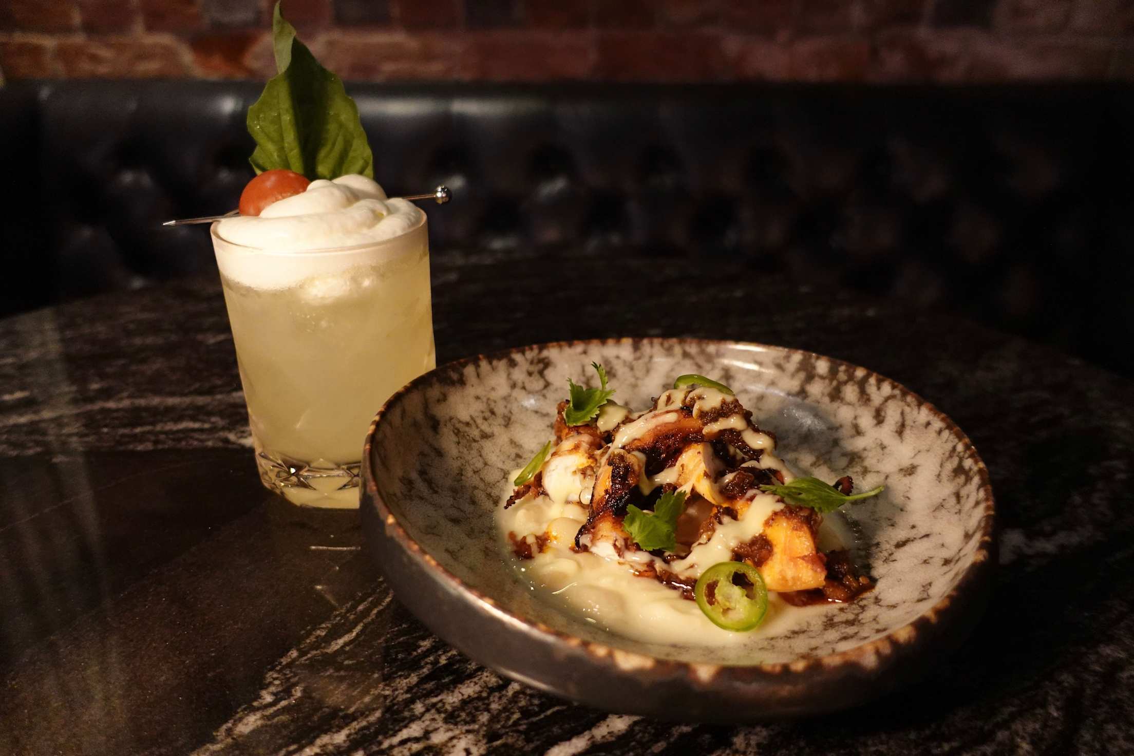 a pale yellow cocktail with foam on top and sauce-covered grilled octopus on a small speckled plate