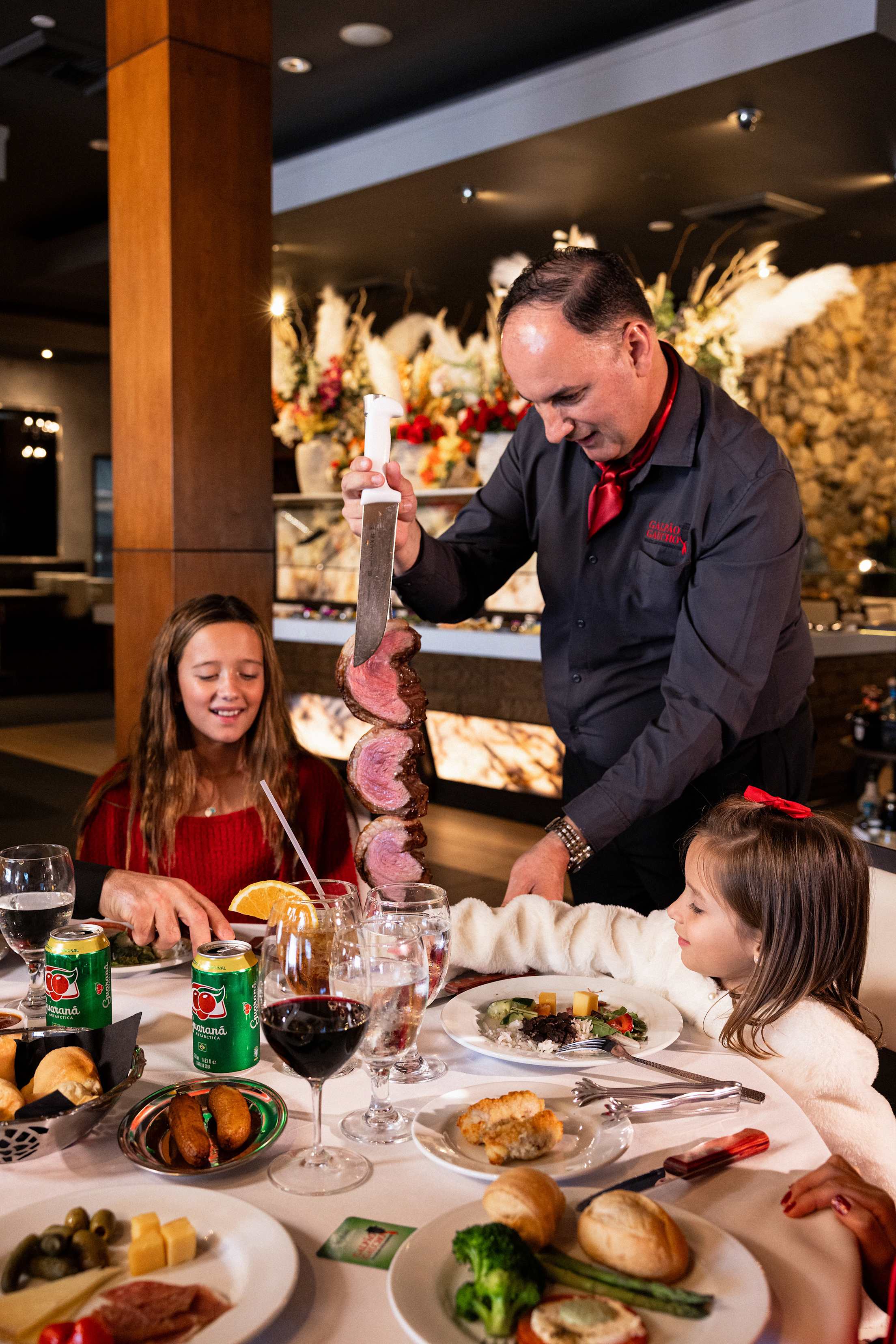 a man cuts steaks at a table full of food while two children sita