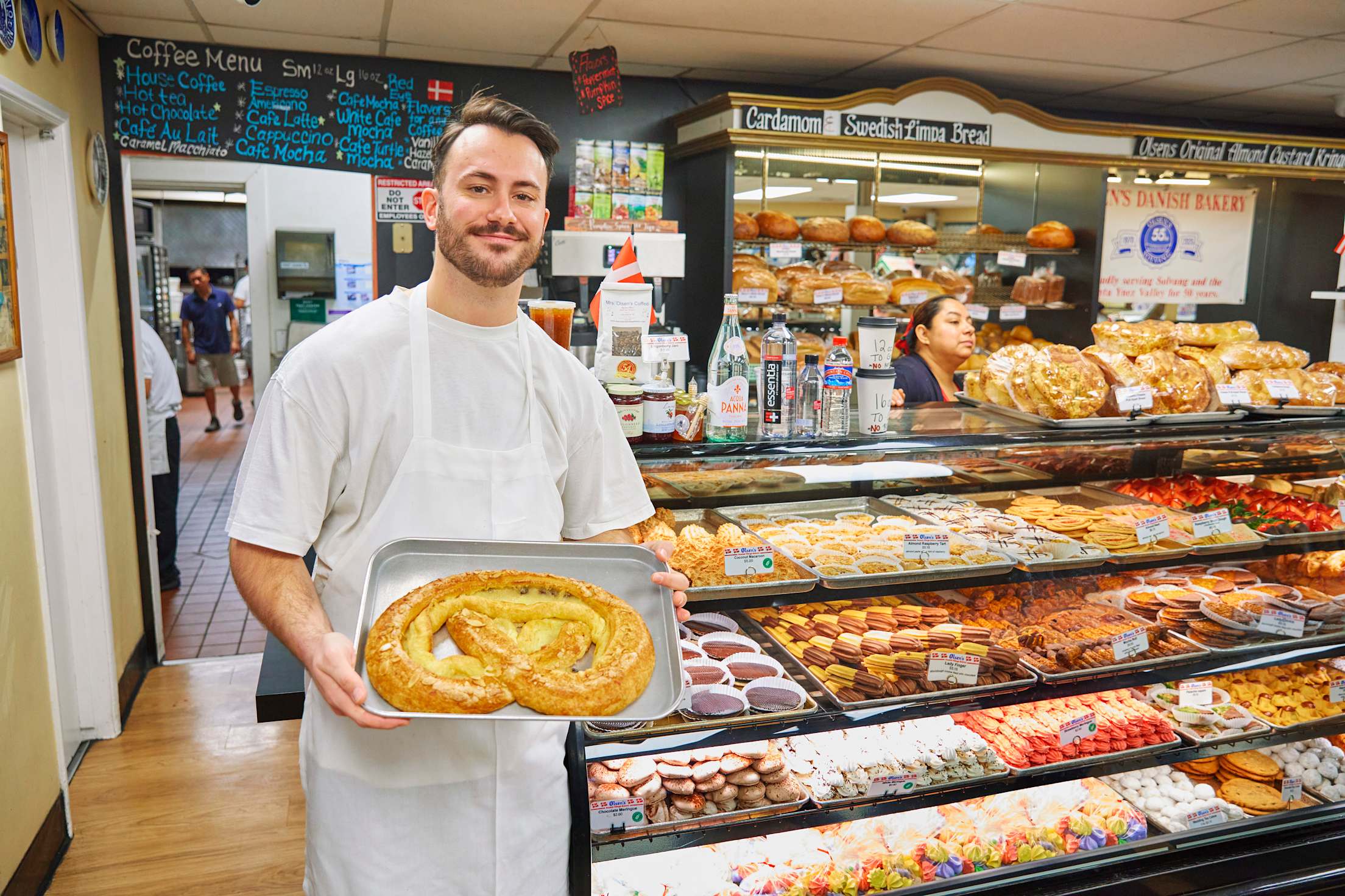 a young white man in a white apron stands holding a giant pastry while standing in front of a pastry display case in a bakery