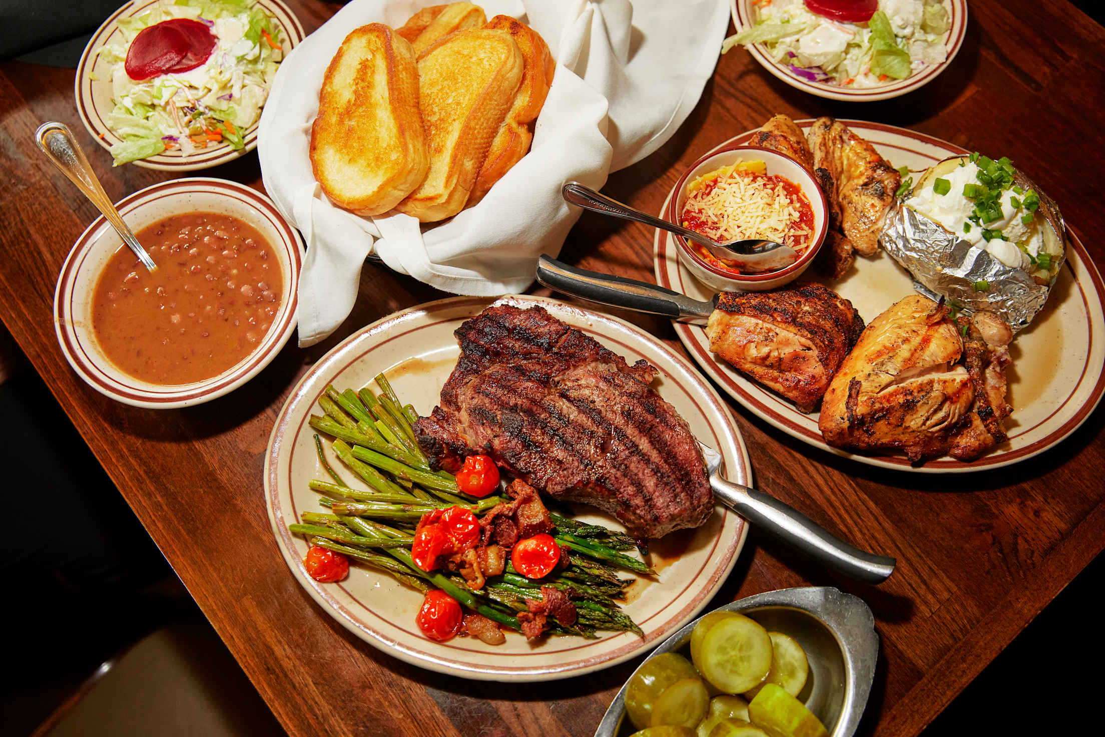 a table covered in dishes: a large steak, garlic bread, asparagus, barbecue chicken, baked potato, beans and pickles