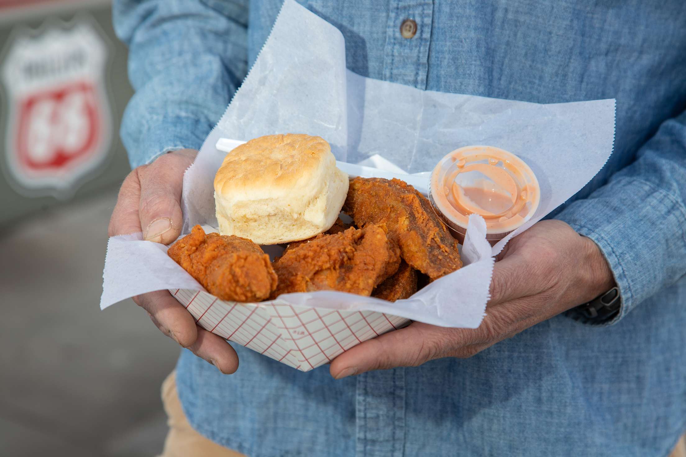 a person in a denim shirt holds a basket with fried chicken, biscuit and sauce
