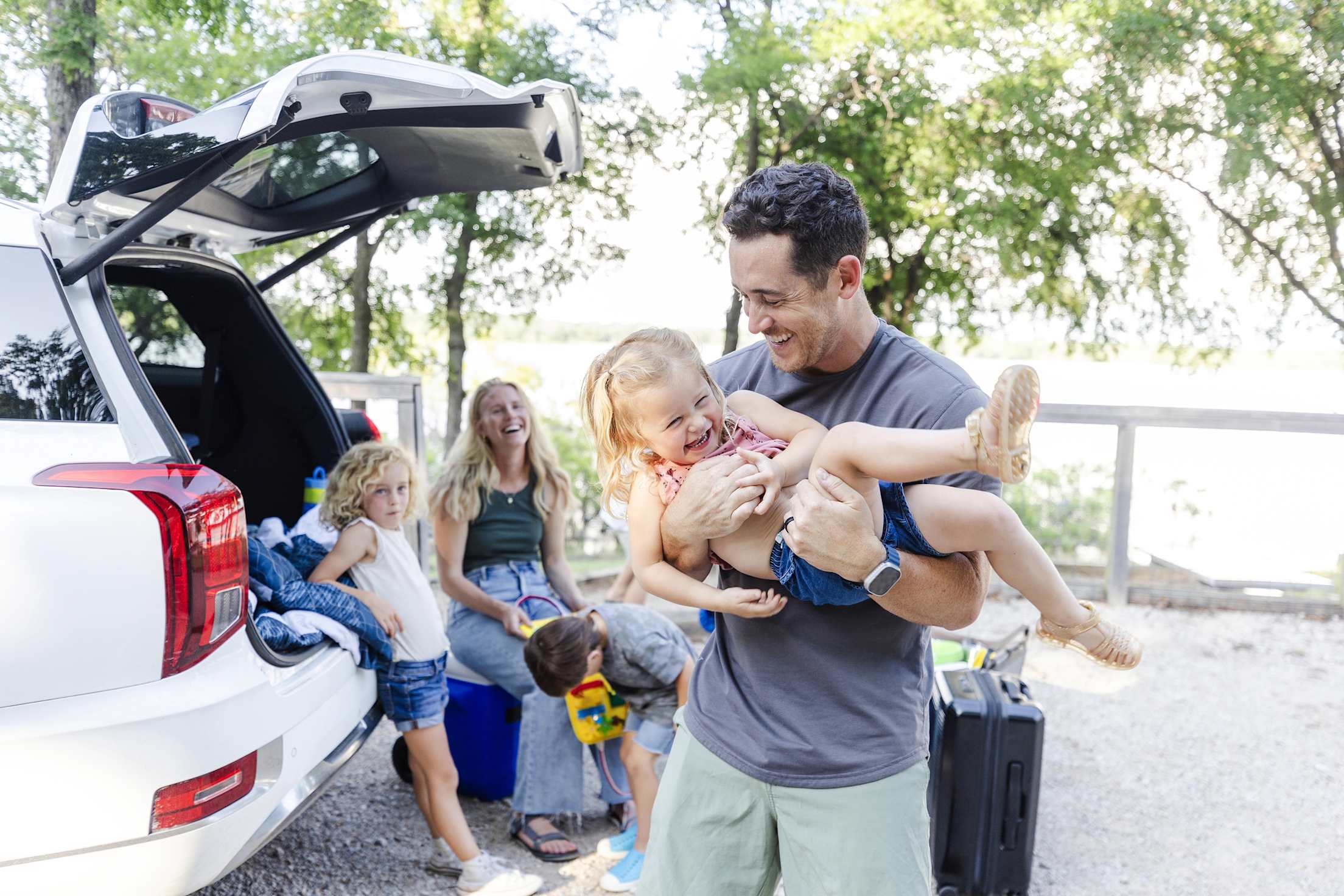 A family laughs and plays together while unloading their luggage from their white SUV's trunk.