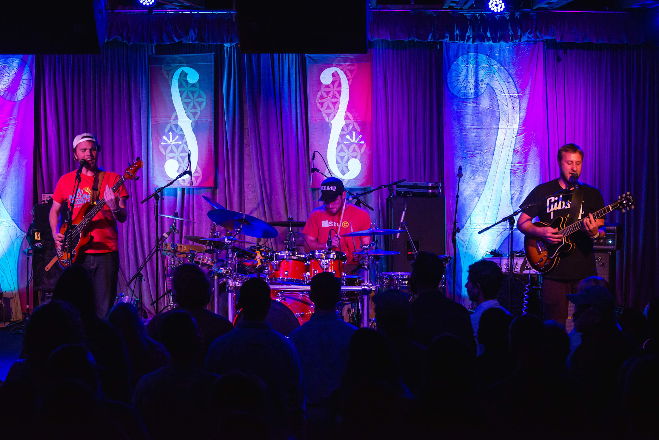 a three piece band plays on a stage lit in purple