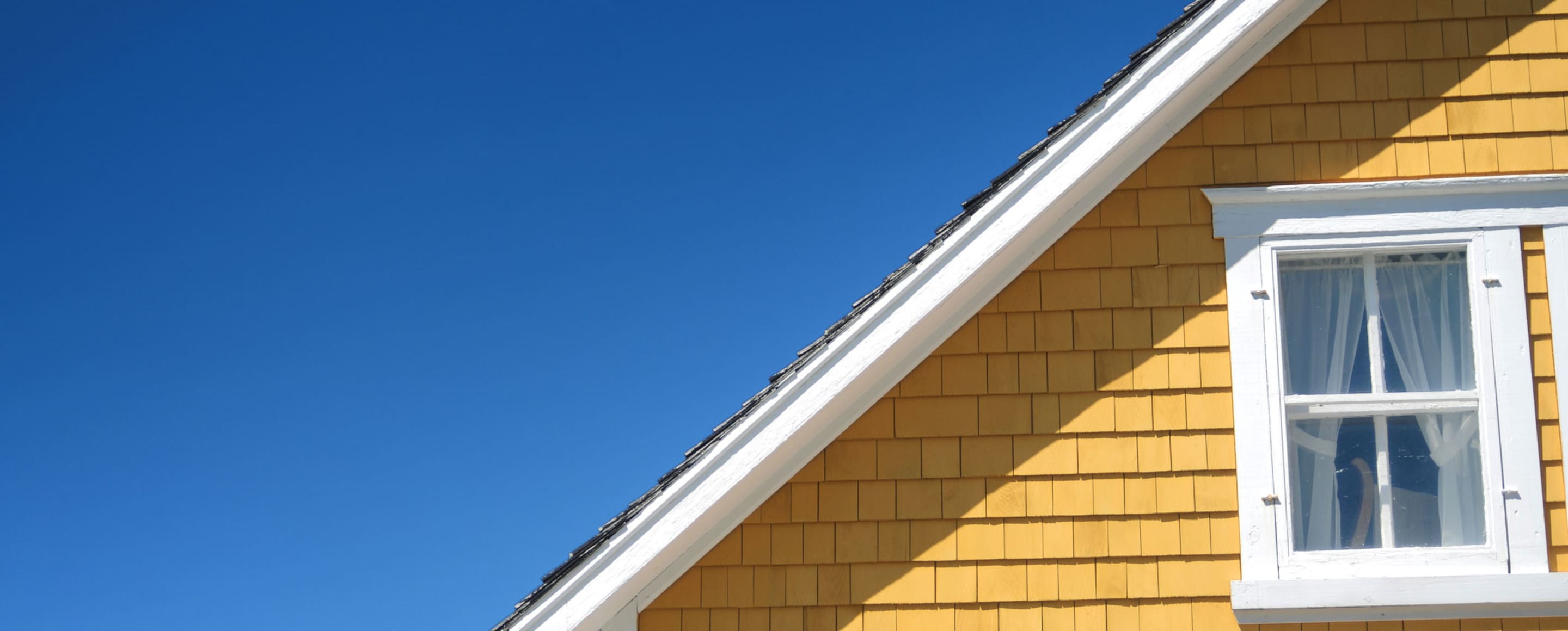 half of the roof of a yellow house with white trim against a bright blue sky.