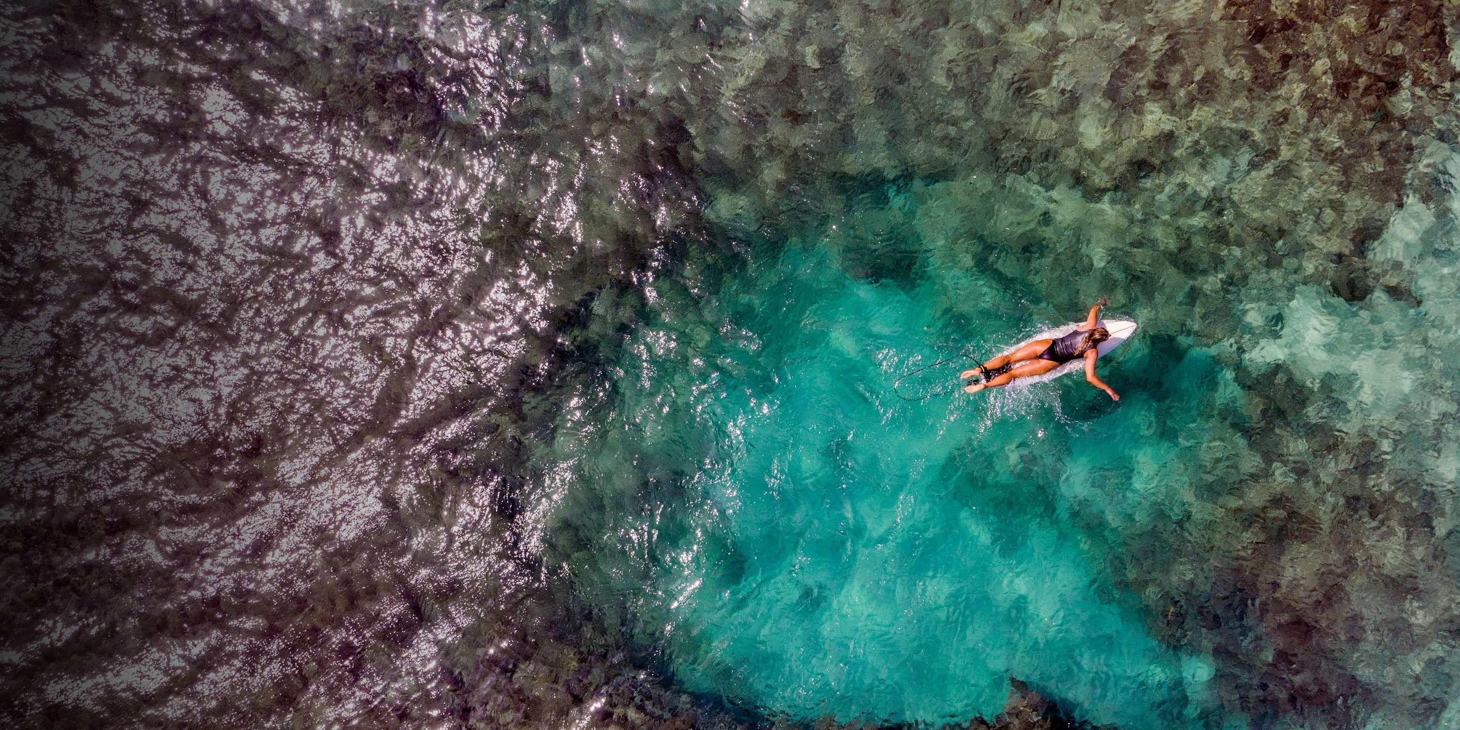 A surfer paddles from a black reef to the ocean