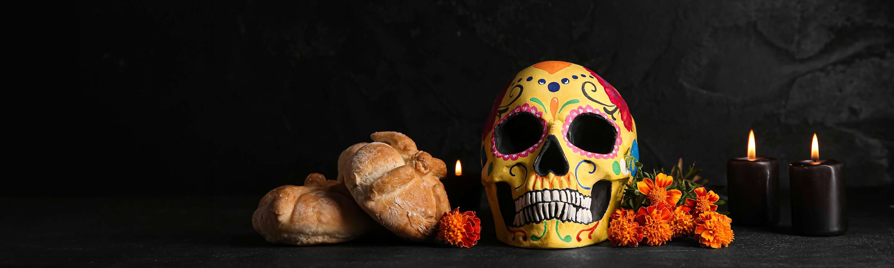 A skull, pan de muerto, and candles on a black table for Dia de los Muertos.