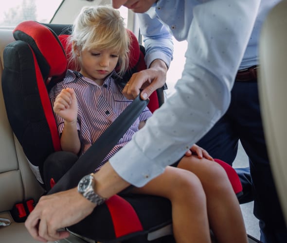 A dad buckles his daughter into her booster seat in the backseat of a sedan.