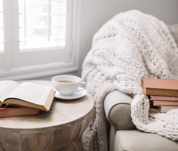 cozy armchair by window with throw, side table, books, cup of tea.