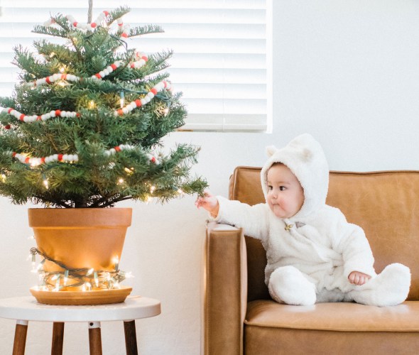A baby on a leather couch reaches out to touch a tabletop Christmas tree.