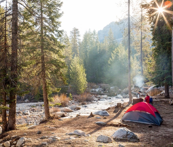 pair of campers with tent pitched beside stream are dressed for cool temps on sunny day at Dorst Creek Campground in Sequoia National Park, California.