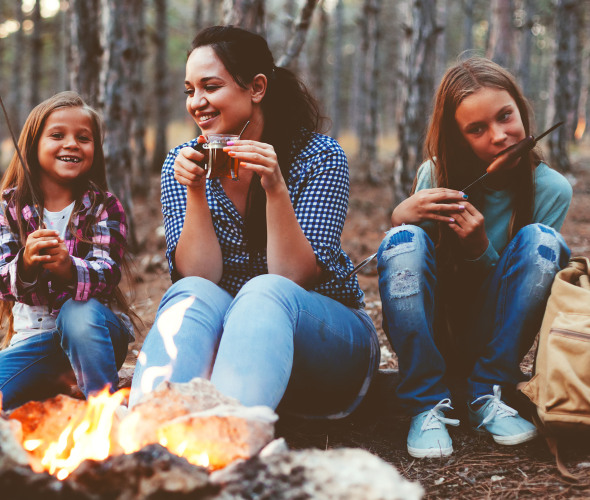 A mom and her two daughters sit around the campfire at a campsite in fall.