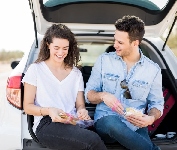A couple sit on the tailgate and at a sandwich on the side of the road.