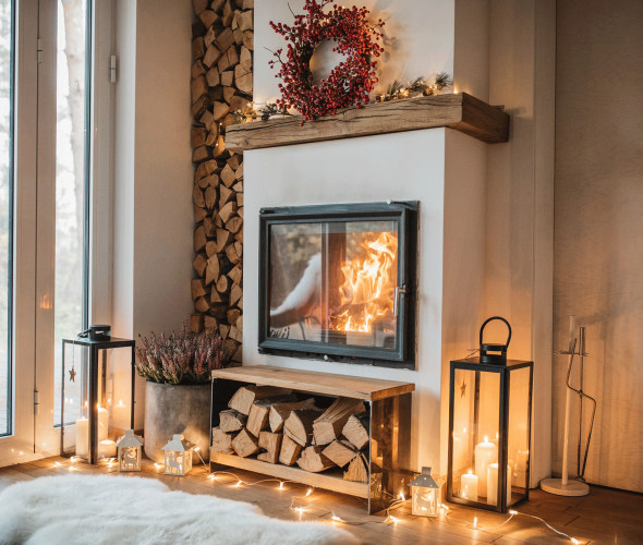 interior with active fireplace, split and stacked firewood, and holiday wreath hung on above on modern mantel beside framed glass door.