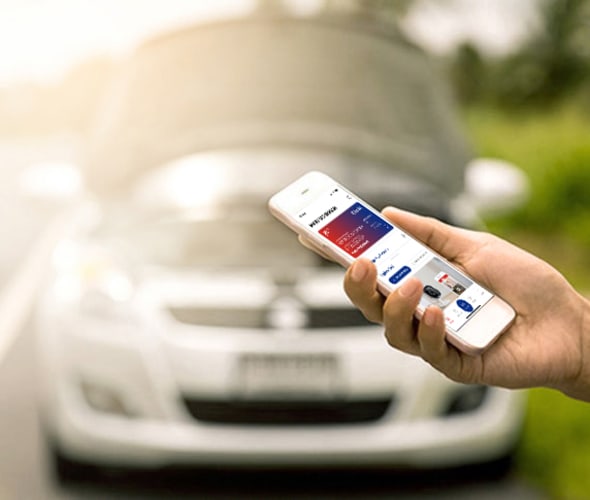 Hand of a AAA Member using the AAA app on their cellphone to call for roadside assistance, with broken down car in the background.