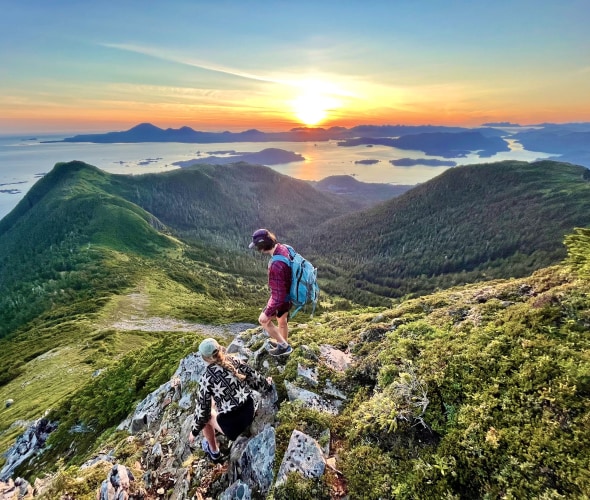 Two hikers scramble on rocks along trail overlooking Sitka sound at sunset.
