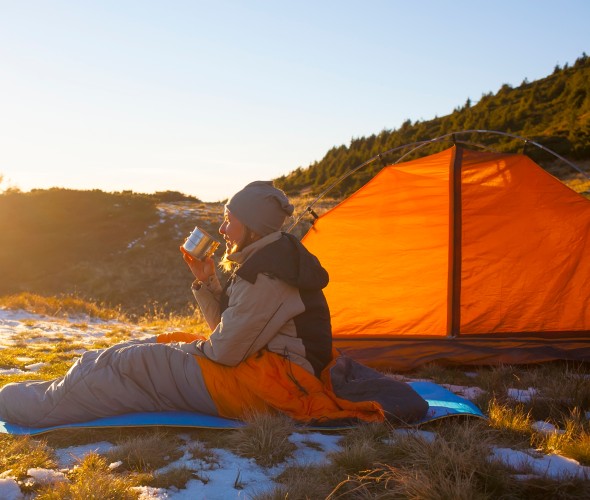 A camper sits on a sleeping pad and sleeping bag on frozen ground while drinking a coffee.