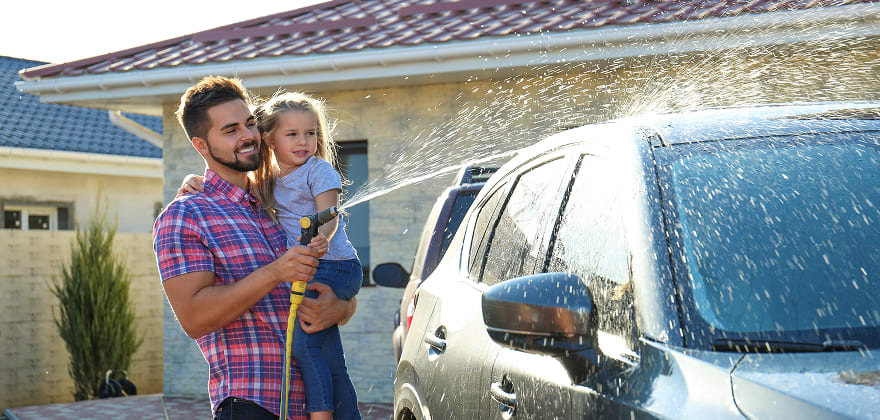 AAA Member covered with AAA Insurance holds child while washing car in front of their house