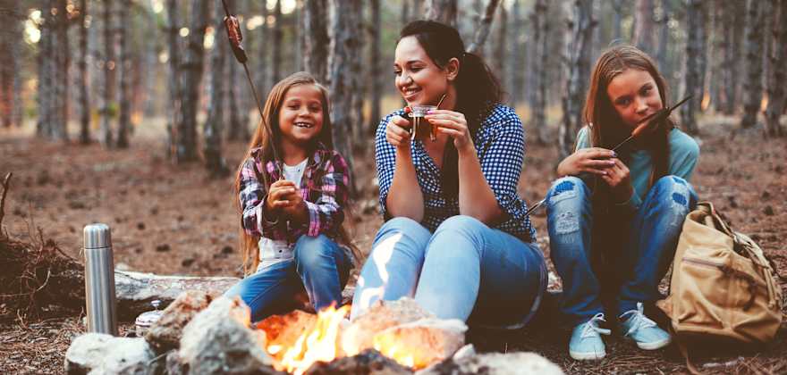 A mom and her two daughters sit around the campfire at a campsite in fall.