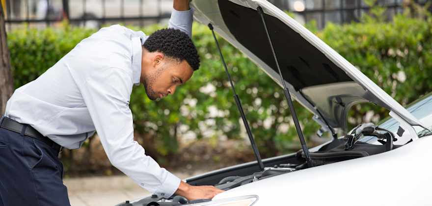 A driver in dress clothes looks under the hood of his white car.
