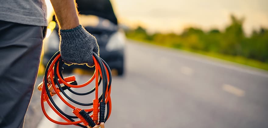 A driver walks toward their car with the hood up with jumper cables.
