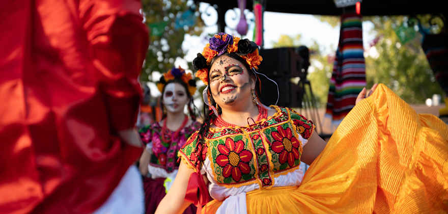 a woman dances in a colorful dress and face paint at a Day of the Dead celebration