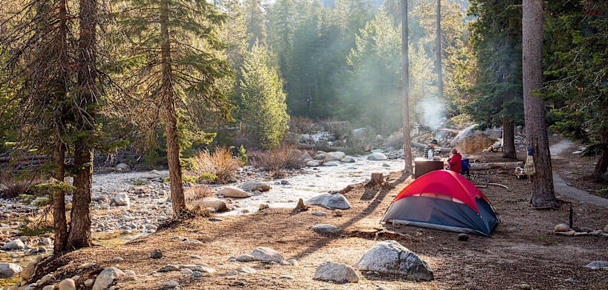 pair of campers with tent pitched beside stream are dressed for cool temps on sunny day at Dorst Creek Campground in Sequoia National Park, California.